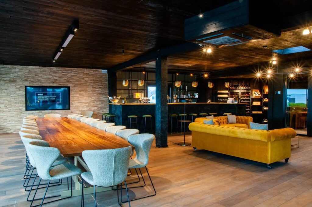 Moderne vergaderruimte met houten tafel, stijlvolle stoelen, gele bank en bar in warme tinten.