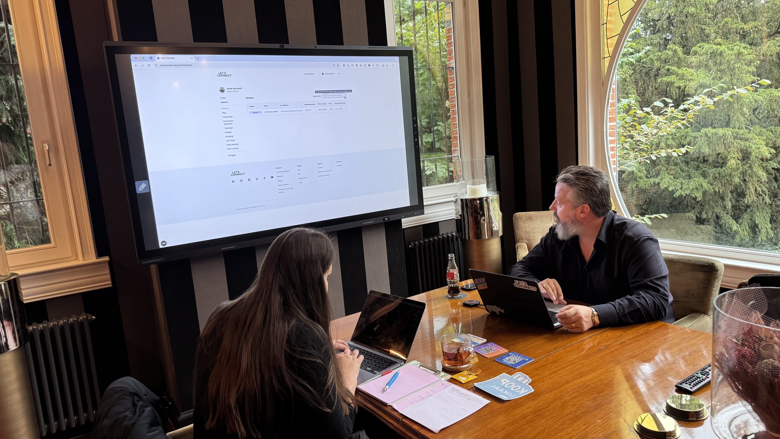 Twee mensen werken aan laptops tijdens een vergadering, met een groot scherm en ramen met uitzicht op bomen.